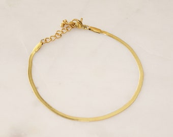 May include: A gold-coloured herringbone chain bracelet. The bracelet has a delicate, flat chain design with an adjustable length. The clasp and connecting hardware are also gold-toned. The bracelet is shown against a light background.