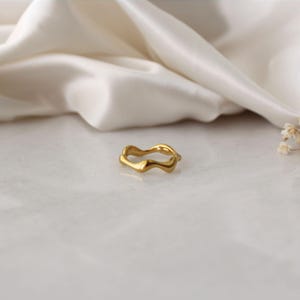 Może przedstawiać: Złoty pierścionek o abstrakcyjnym, falistym wzorze. Pierścionek wykonany jest z metalu i ma gładkie, polerowane wykończenie.