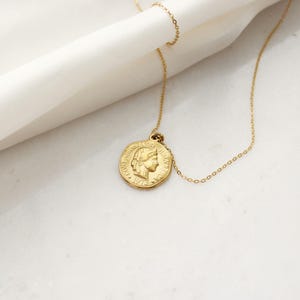 Pode incluir: Um colar com pingente de moeda dourada. O pingente apresenta um perfil e o ano de 1973. A corrente &eacute; fina e dourada. O colar &eacute; exibido em uma superf&iacute;cie branca.