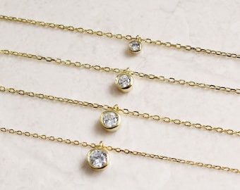 May include: Four gold necklaces with a single round clear stone pendant on each. The necklaces are layered on a white marble surface.