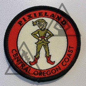 May include: A circular embroidered patch with a black border, a red outer ring, and a cream-colored center. The text PIXIELAND and CENTRAL OREGON COAST is stitched in black. A cartoon elf in a green outfit and red boots stands in the center.