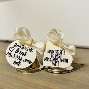 May include: Two silver bells with white satin bows and cream-colored tags. The tags have black text that reads "Ring the Bell to wish Mr. & Mrs. Jones well" and "Ring the Bell to wish Mr & Mrs. Smith well".