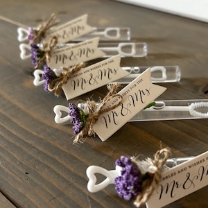 May include: Wedding favor bubble wands with a heart-shaped wand and a small bouquet of purple flowers. The wands are decorated with a tag that says "Bubbles and Wishes for the Mr & Mrs".