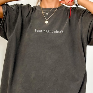 Peut inclure: T-shirt gris foncé surdimensionné avec le texte "team night shift" brodé en blanc. Le t-shirt a des manches courtes et une coupe décontractée. Le tissu semble être une matière douce et lavée.