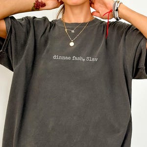 May include: Dark grey oversized t-shirt with short sleeves. The shirt features the text "dinnae fash, Slav" in a small, centered font. The shirt has a slightly faded appearance.