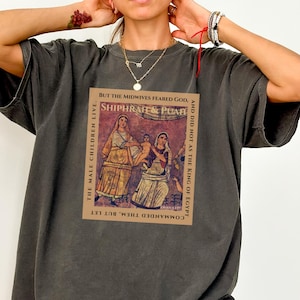 May include: Dark grey t-shirt with a rectangular graphic featuring the text "SHIPHRAH & PUAH" and other text. The graphic shows two women in ancient clothing. The shirt has short sleeves and a loose fit.