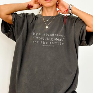 以下が含まれることがあります： 「My Husband is out 'Providing Meat' for the family」のテキストが入った濃いグレーのTシャツ。Tシャツは半袖でゆったりとしたフィット感。テキストはシンプルなサンセリフ体です。