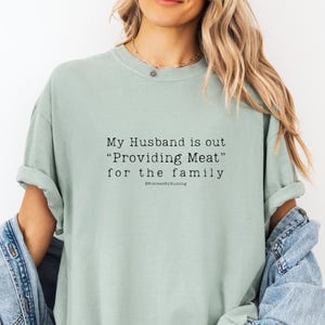 以下が含まれることがあります： 「My Husband is out 'Providing Meat' for the family」の黒い文字が入った、薄緑色のTシャツ。 ゆったりとしたフィット感で、袖はロールアップされています。 テキストはシャツの前面の中央に配置されています。