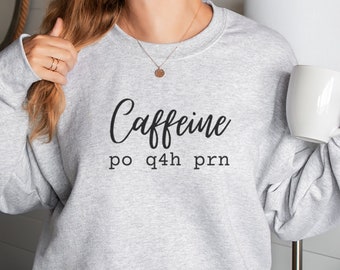 Op de afbeelding: Een grijs sweatshirt met de tekst "Caffeine po q4h prn" in zwarte letters. Het sweatshirt wordt gedragen door een persoon. Het woord "Ash" is zichtbaar in de rechterbenedenhoek van de afbeelding.