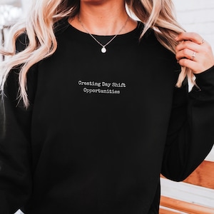 Peut inclure: Sweat-shirt noir avec le texte "Creating Day Shift Opportunities" en blanc. Le sweat-shirt a des manches longues. Le modèle porte un collier argenté. Le tissu semble doux.