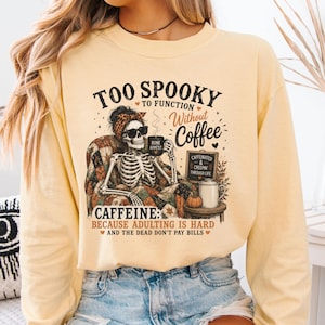 May include: A pale yellow long-sleeve sweatshirt featuring a graphic of a skeleton wearing a leopard print headband and sunglasses, sitting in a quilt-covered chair with a coffee mug. Text reads: TOO SPOOKY TO FUNCTION WITHOUT COFFEE CAFFEINE: BECAUSE ADULTING IS HARD AND THE DEAD DON'T PAY BILLS.