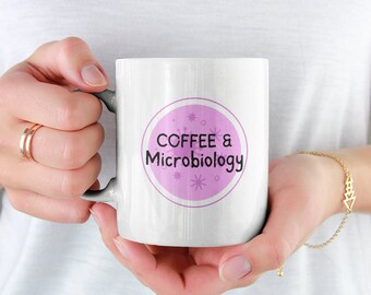 May include: White ceramic mug with a pink circle design. The circle has the text "COFFEE & Microbiology" in black text with small white stars and circles around the text.