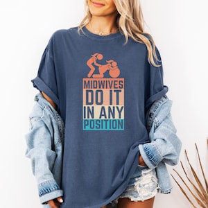 Könnte beinhalten: Ein marineblaues T-Shirt mit einer Grafik im Vintage-Stil, auf dem "Midwives Do It In Any Position" steht. Die Grafik zeigt eine Strichmännchen-Illustration einer Hebamme, die eine Person bei der Geburt unterstützt.
