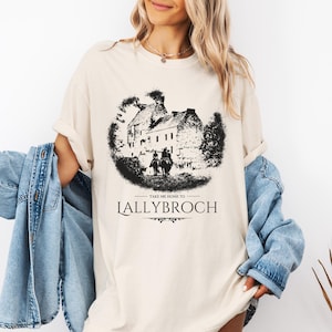 May include: A beige t-shirt featuring a black and white illustration of a castle and two people riding horses. The text "Take me home to Lallybroch" is printed below the illustration.