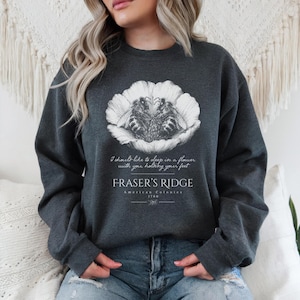 Puede incluir: Una sudadera de cuello redondo en color gris carbón oscuro con un gráfico blanco de dos abejas dentro de una flor. El texto dice: I should like to sleep in a flower with you, holding your feet. FRASER'S RIDGE American Colonies 1780, con un pequeño icono de abeja.