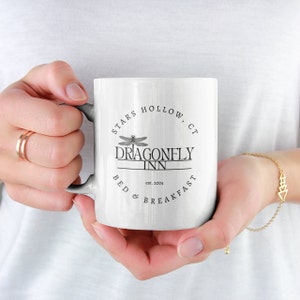 Può includere: Tazza in ceramica bianca con una grafica in bianco e nero di una libellula e il testo "Stars Hollow, CT Dragonfly Inn est. 2004 Bed & Breakfast".
