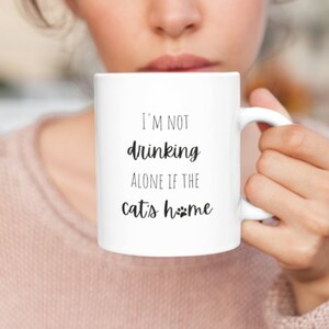 Puede incluir: Taza de café de cerámica blanca con texto negro que dice "No estoy bebiendo solo si el gato está en casa" con un icono de huella de pata.
