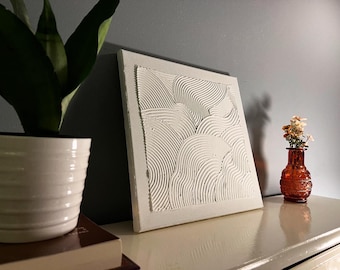 May include: A white textured artwork with a wave design, displayed on a white surface. A potted plant and a small orange vase with flowers are also present. The square artwork has a raised, textured surface.