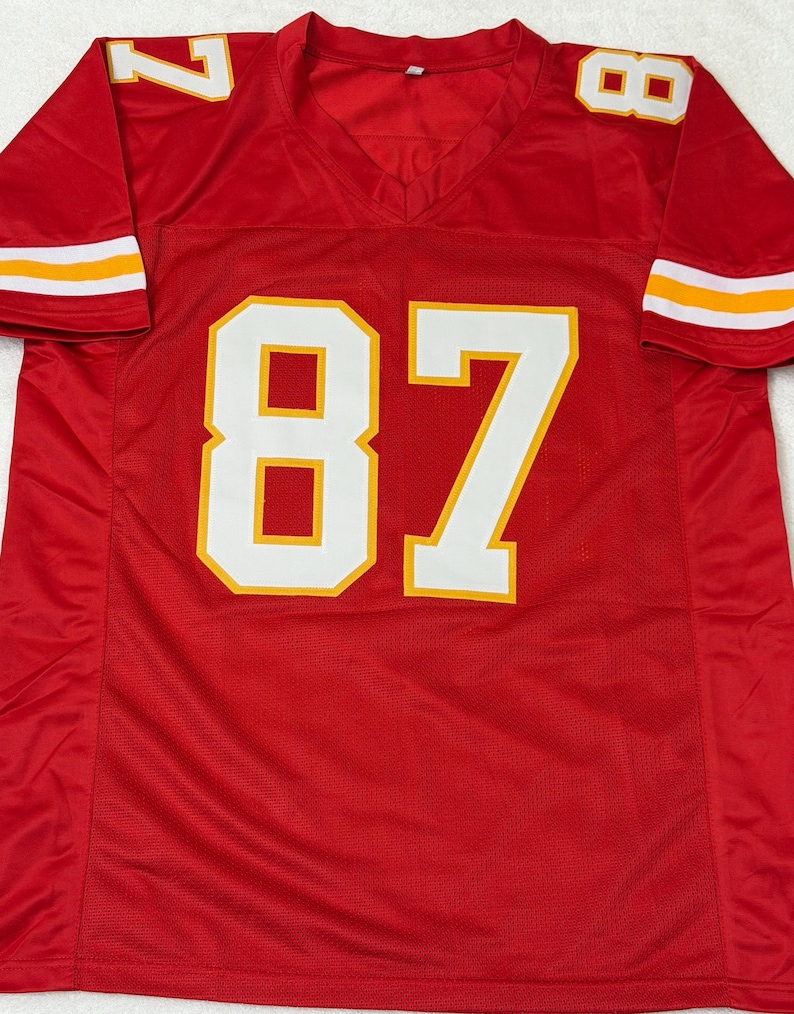 May include: A red football jersey with the number 87 in white with a yellow outline on the front. The number 7 is on the left shoulder and the number 8 is on the right shoulder. The sleeves have white and yellow stripes.