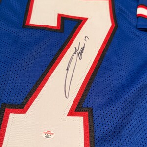 Puede incluir: Camiseta de malla azul con un gran n&uacute;mero 7 blanco delineado en negro y rojo. El n&uacute;mero est&aacute; firmado con tinta negra. Una peque&ntilde;a pegatina blanca con texto negro es visible.