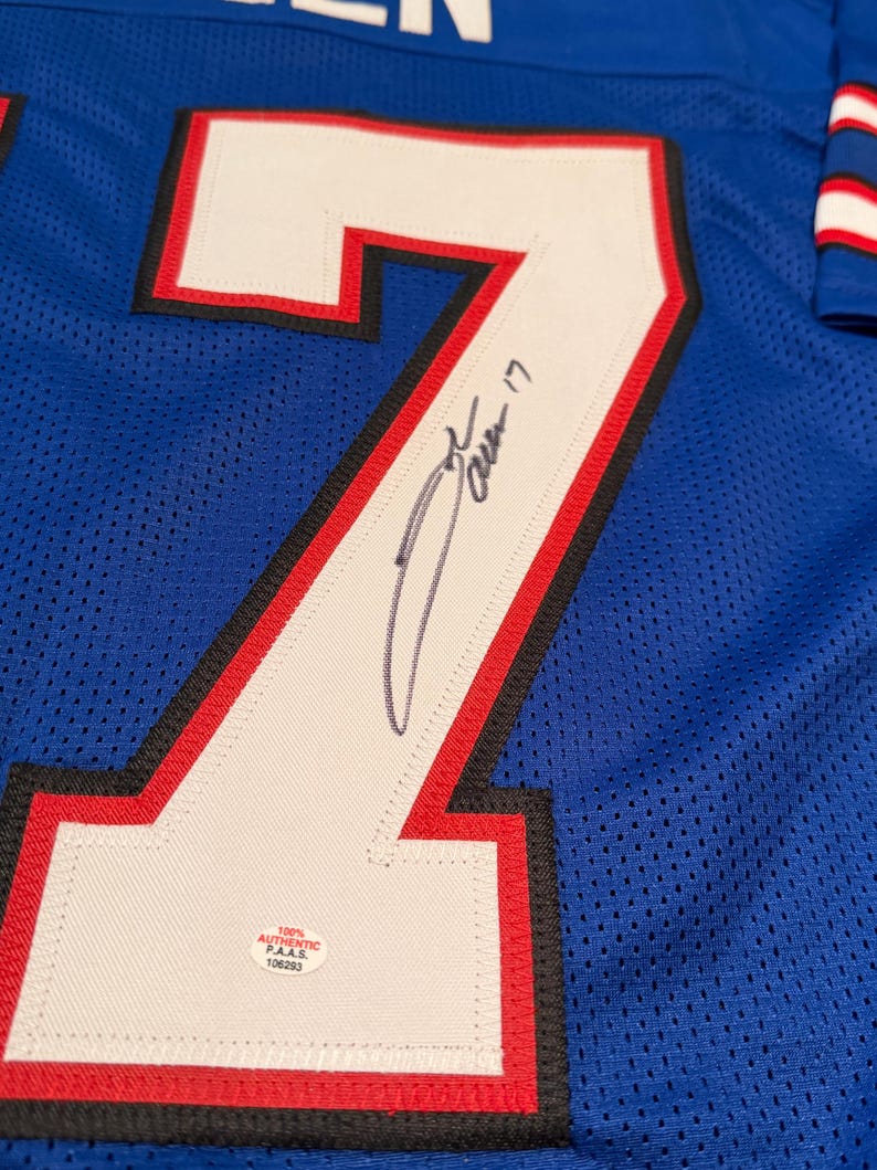 Puede incluir: Camiseta de malla azul con un gran n&uacute;mero 7 blanco delineado en negro y rojo. El n&uacute;mero est&aacute; firmado con tinta negra. Una peque&ntilde;a pegatina blanca con texto negro es visible.