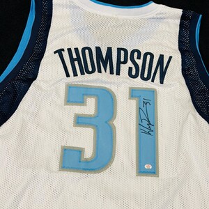 May include: White mesh jersey with the name "THOMPSON" and the number "31" in light blue and navy blue. The jersey features a signature and a PSA/DNA authentication sticker.