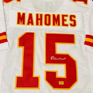 Puede incluir: Una camiseta deportiva blanca con el nombre "MAHOMES" en letras rojas con bordes amarillos en la parte posterior. El n&uacute;mero "15" aparece en grandes d&iacute;gitos rojos y amarillos debajo del nombre, junto a una firma plateada y una peque&ntilde;a pegatina de autenticaci&oacute;n dorada.