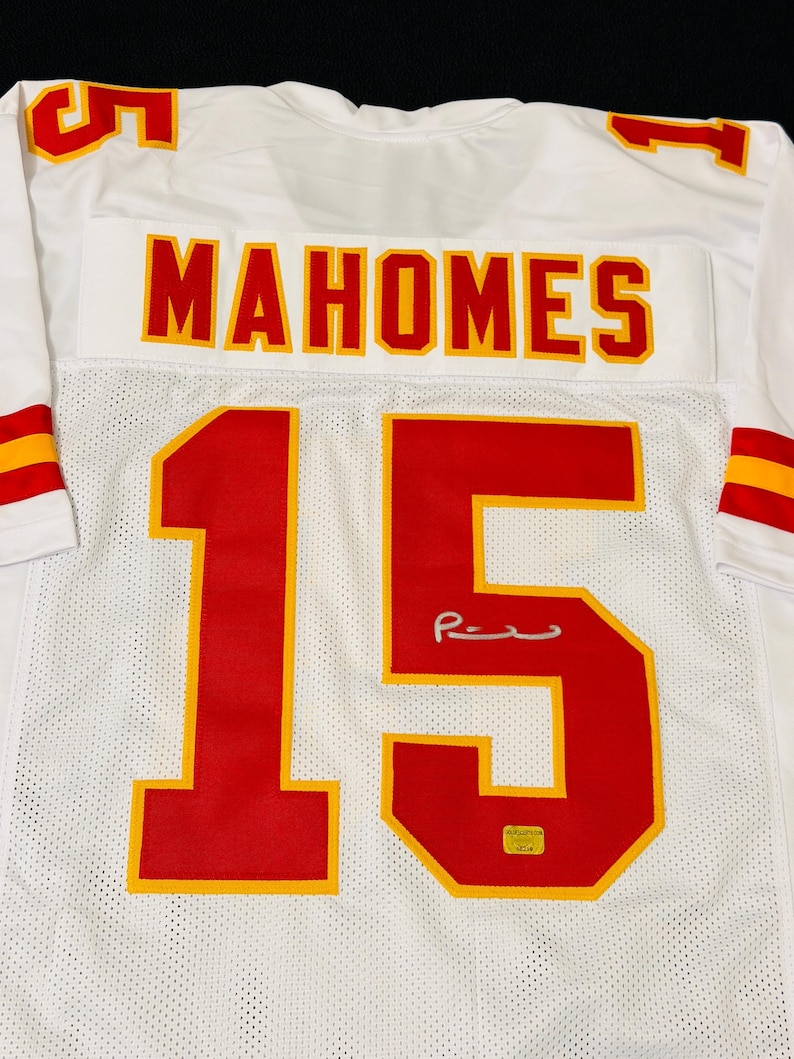 Puede incluir: Una camiseta deportiva blanca con el nombre "MAHOMES" en letras rojas con bordes amarillos en la parte posterior. El n&uacute;mero "15" aparece en grandes d&iacute;gitos rojos y amarillos debajo del nombre, junto a una firma plateada y una peque&ntilde;a pegatina de autenticaci&oacute;n dorada.