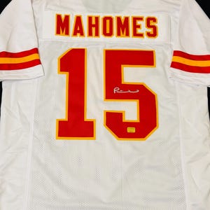 Puede incluir: Una camiseta deportiva blanca con el nombre MAHOMES y el n&uacute;mero 15 en rojo con bordes amarillos en la parte posterior. Las mangas tienen rayas rojas y amarillas, y una peque&ntilde;a firma es visible en el n&uacute;mero 15, junto con una peque&ntilde;a pegatina amarilla.
