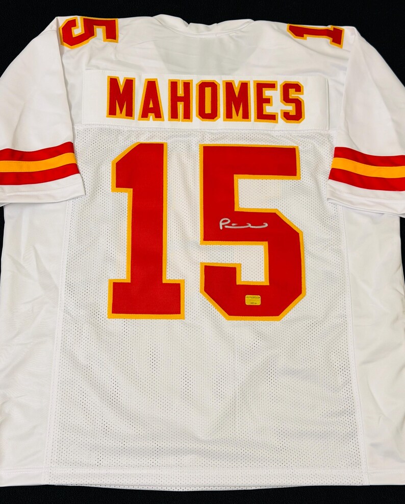 Puede incluir: Una camiseta deportiva blanca con el nombre MAHOMES y el n&uacute;mero 15 en rojo con bordes amarillos en la parte posterior. Las mangas tienen rayas rojas y amarillas, y una peque&ntilde;a firma es visible en el n&uacute;mero 15, junto con una peque&ntilde;a pegatina amarilla.