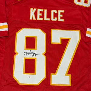 May include: Red football jersey with white and gold accents. The jersey features the name "KELCE" across the back and the number "87" in large white numerals with gold trim. The jersey is signed with a black marker.