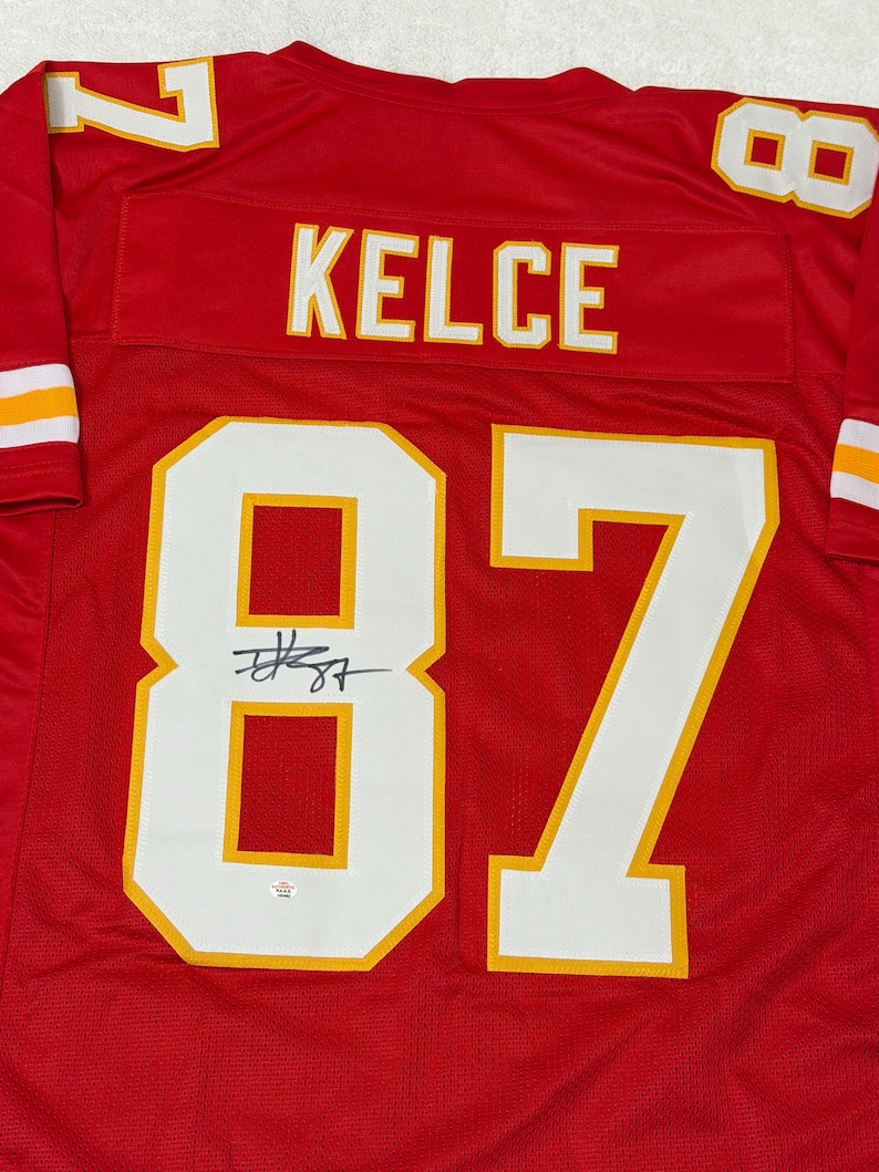 May include: Red football jersey with white and gold accents. The jersey features the name "KELCE" across the back and the number "87" in large white numerals with gold trim. The jersey is signed with a black marker.