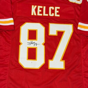 May include: A red football jersey with white and yellow accents. The jersey features the name "KELCE" across the back and the number "87" in large white numerals with yellow trim. The number "7" is visible on the shoulders.
