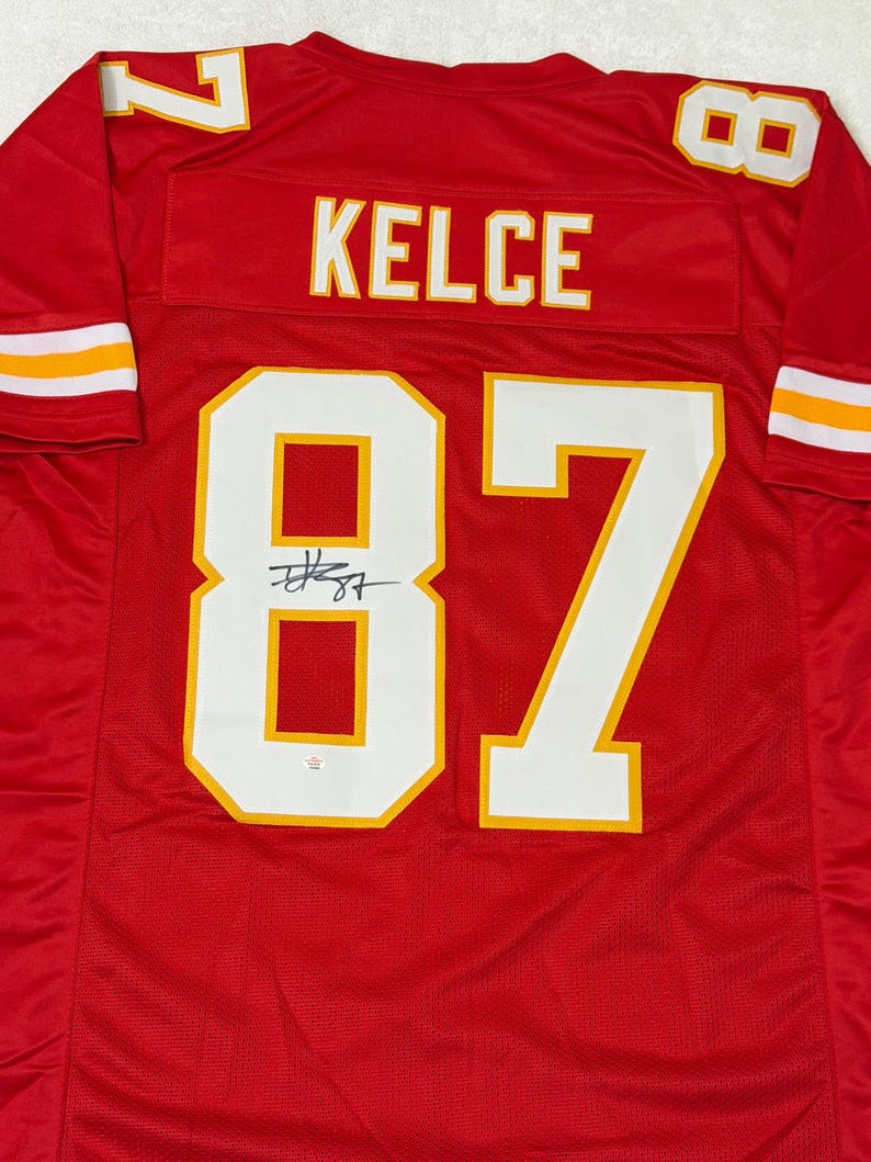 May include: A red football jersey with white and yellow accents. The jersey features the name "KELCE" across the back and the number "87" in large white numerals with yellow trim. The number "7" is visible on the shoulders.