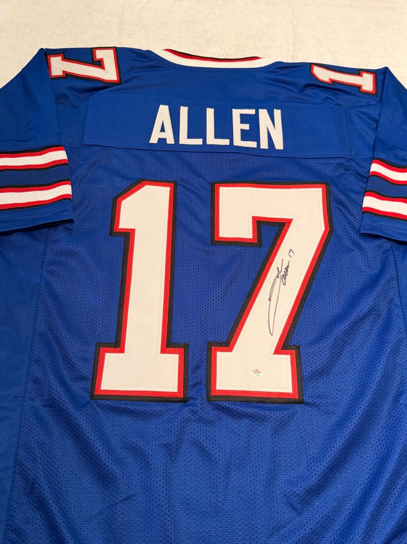 Puede incluir: Una camiseta de f&uacute;tbol azul con el nombre "ALLEN" en la espalda y el n&uacute;mero "17" en blanco con ribetes rojos y negros. La camiseta tiene rayas blancas y rojas en las mangas y una firma en el n&uacute;mero.