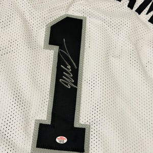 以下が含まれることがあります： 白いメッシュのバスケットボールジャージ。グレーの縁取りが施された大きな黒い数字の「1」が中央にあり、銀色のインクでサインが書かれています。数字の下部には「100% AUTHENTIC P.A.A.S. 106136」と書かれた小さな円形のステッカーが貼られています。名前の一部が見えています。