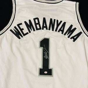 以下が含まれることがあります： 背面上部に黒い文字でWEMBANYAMAと書かれた白いバスケットボールジャージ。名前の下には、グレーの縁取りが施された黒い数字の1がある。数字の上には黒いサインと小さな円形のステッカーが見える。