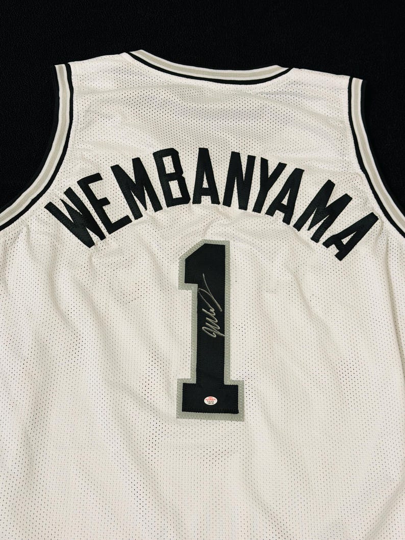 以下が含まれることがあります： 背面上部に黒い文字でWEMBANYAMAと書かれた白いバスケットボールジャージ。名前の下には、グレーの縁取りが施された黒い数字の1がある。数字の上には黒いサインと小さな円形のステッカーが見える。