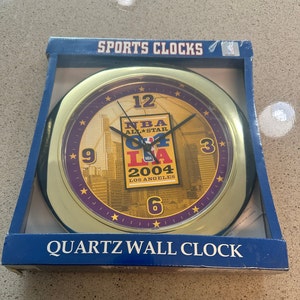 May include: A gold-toned quartz wall clock featuring the NBA All-Star 2004 Los Angeles logo. The clock face has a cityscape design and a purple ring with star accents. The clock is in its original blue box, labeled "Sports Clocks."