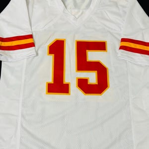 Puede incluir: Una camiseta deportiva blanca de manga corta con el n&uacute;mero 15 en rojo con un borde amarillo en la parte delantera. Las mangas tienen dos rayas horizontales rojas y amarillas, y el n&uacute;mero 15 tambi&eacute;n est&aacute; impreso en rojo en cada hombro.