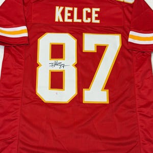 May include: Red football jersey with the name "KELCE" and the number "87" in white with yellow trim. The jersey has a signature below the number. The sleeves have white and yellow stripes.