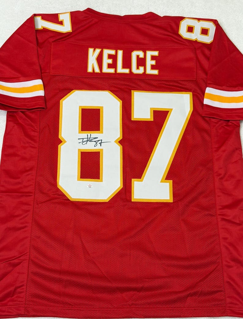 May include: Red football jersey with the name "KELCE" and the number "87" in white with yellow trim. The jersey has a signature below the number. The sleeves have white and yellow stripes.