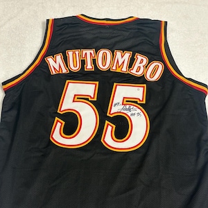 Può includere: Maglia da basket nera con il nome "MUTOMBO" in lettere rosse e oro. Il numero "55" è visualizzato in bianco con un contorno rosso e oro. La maglia ha una firma sopra il numero. Rifiniture rosse e oro.