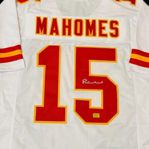 Puede incluir: Una camiseta deportiva blanca con el nombre MAHOMES y el n&uacute;mero 15 en rojo con bordes amarillos en la parte posterior. La camiseta incluye rayas rojas y amarillas en las mangas y una firma sobre el n&uacute;mero 15 con una peque&ntilde;a etiqueta de autenticidad.