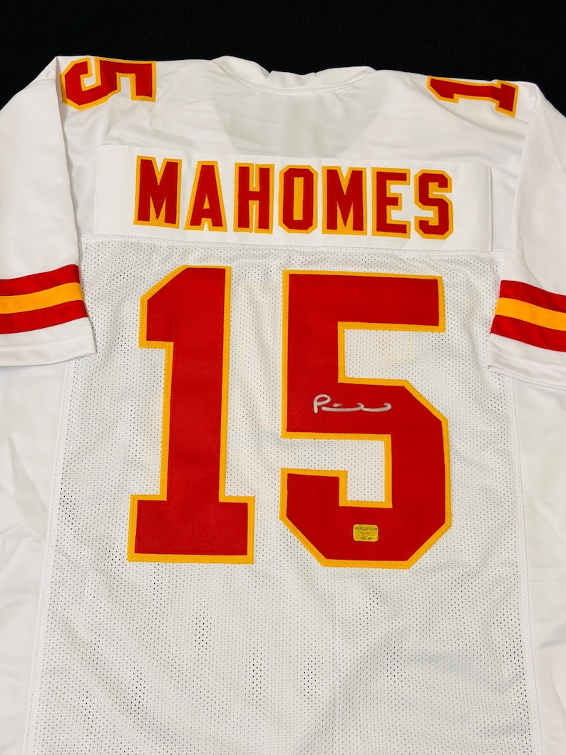 Puede incluir: Una camiseta deportiva blanca con el nombre MAHOMES y el n&uacute;mero 15 en rojo con bordes amarillos en la parte posterior. La camiseta incluye rayas rojas y amarillas en las mangas y una firma sobre el n&uacute;mero 15 con una peque&ntilde;a etiqueta de autenticidad.