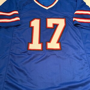 Puede incluir: Una camiseta de malla azul con el n&uacute;mero 17 en blanco con ribetes rojos y negros. La camiseta tiene rayas blancas y rojas en las mangas y un cuello en V con ribetes blancos y rojos.