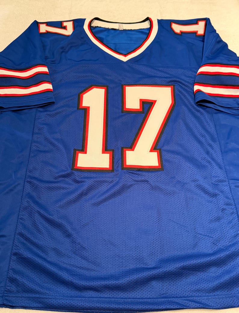 Puede incluir: Una camiseta de malla azul con el n&uacute;mero 17 en blanco con ribetes rojos y negros. La camiseta tiene rayas blancas y rojas en las mangas y un cuello en V con ribetes blancos y rojos.