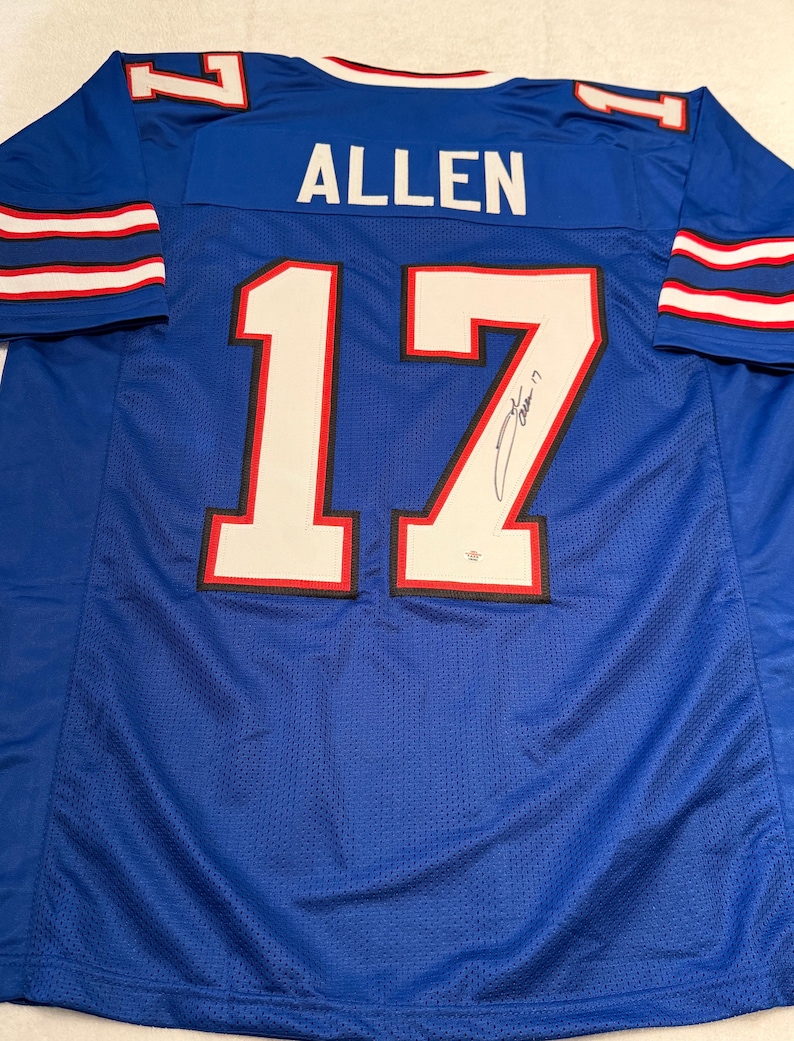 Puede incluir: Una camiseta de f&uacute;tbol azul con el nombre "ALLEN" y el n&uacute;mero "17" en blanco. La camiseta tiene rayas rojas y blancas en las mangas y el n&uacute;mero "17" est&aacute; delineado en rojo. La camiseta parece estar firmada.