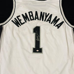 以下が含まれることがあります： 黒いトリム、黒い文字でアーチ状に配置されたWEMBANYAMAの名前、シルバーの縁取りが施された黒い背番号1が特徴の白いバスケットボールジャージ。背番号1には黒いインクのサインと小さな円形の認証ステッカーが貼られている。