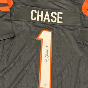 Puede incluir: Una camiseta de fútbol gris con detalles en naranja en las mangas. El nombre "CHASE" está impreso en blanco en los hombros. El número "1" está en la parte delantera, con una firma encima. La camiseta está hecha de un material de malla transpirable.
